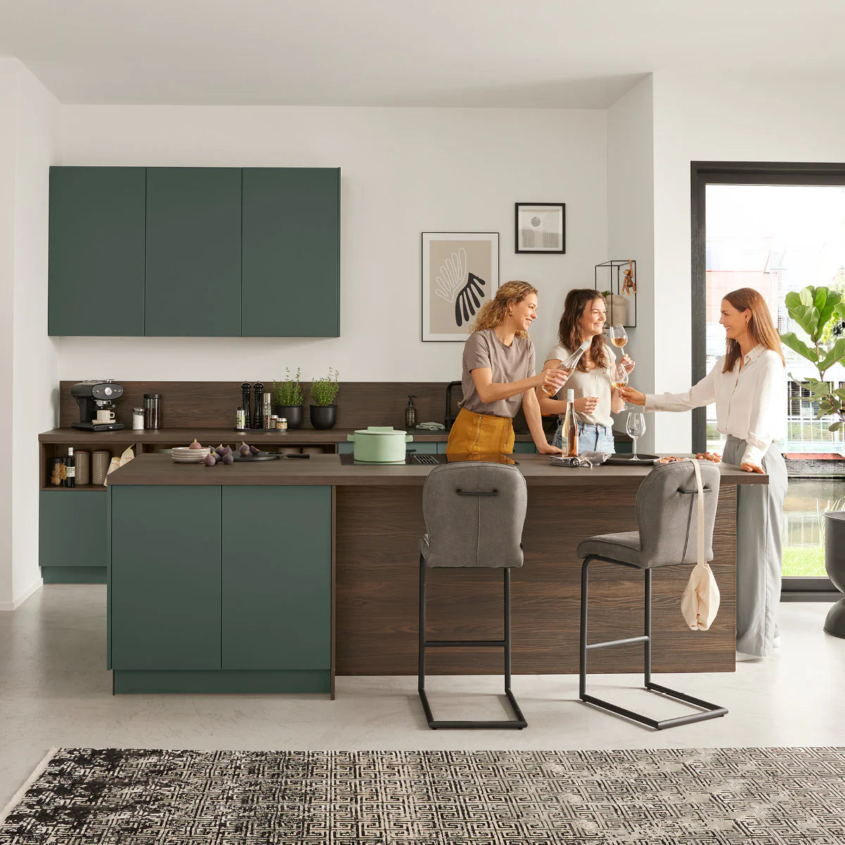 Interliving Küche mit dunkelgrüner Front und Nussbaum-Optik, Frauen stoßen an der Kochinsel an – Interliving Lieblingsstücke bei FREY