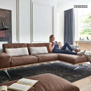Braunes Ledersofa von Koinor mit moderner Form, auf dem eine Frau entspannt sitzt – wohnliches Ambiente mit Fenster und Teppich.
