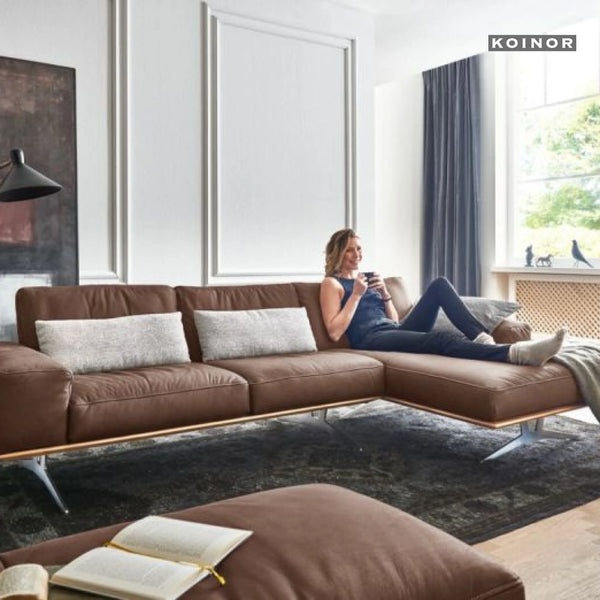 Braunes Ledersofa von Koinor mit moderner Form, auf dem eine Frau entspannt sitzt – wohnliches Ambiente mit Fenster und Teppich.